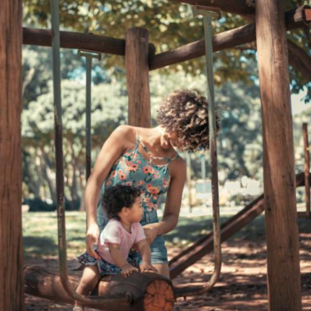 Uma mãe brinca com sua filha em um parquinho, cercadas por árvores e luz natural.
